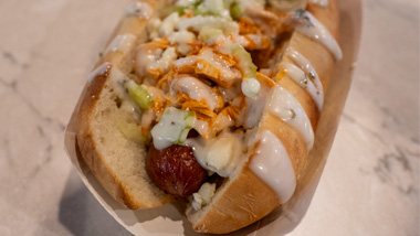 Close-up of a Zombie Dogz hot dog from Plainridge Park Casino, featuring a grilled hot dog in a bun topped with shredded buffalo chicken, blue cheese crumbles, and a white creamy sauce.